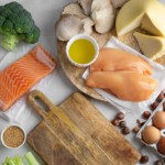 A flatlay of Mediterranean Diet staples including salmon, chicken, eggs, broccoli, cheese, and olive oil. A wooden cutting board is in the center of the scene. 