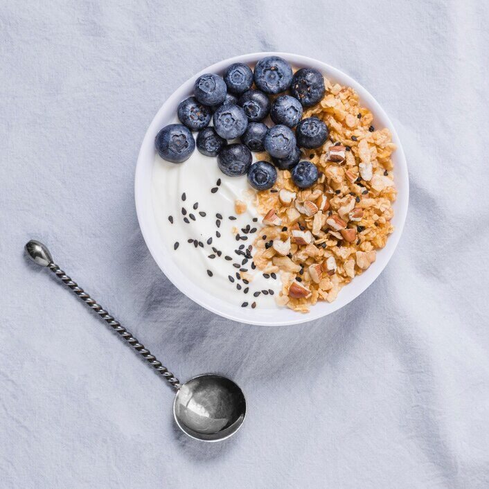 A white bowl of yogurt with granola, black sesame seeds, and blueberries, sitting on a light gray tablecloth, next to a silver spoon.