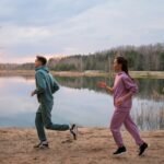 A man and a woman in athletic wear run along the edge of a lake, enjoying a healthy outdoor workout that complements a Mediterranean Diet. 