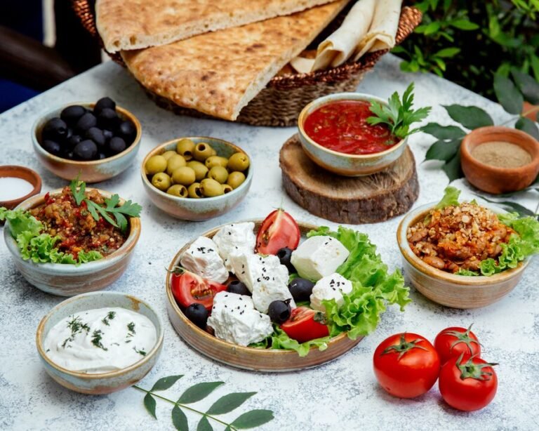 A white table with a variety of Middle Eastern appetizers, such as a tomato and feta salad, baba ghanoush, a lentil salad, and some olives, as well as a basket of flatbreads and a small bowl of tomato sauce.