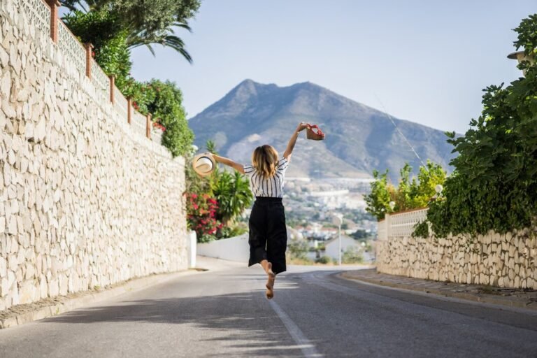 A woman in a striped shirt and black pants jumps on a street with a mountain backdrop, her arms outstretched. She is barefoot and holding a hat in her right hand.