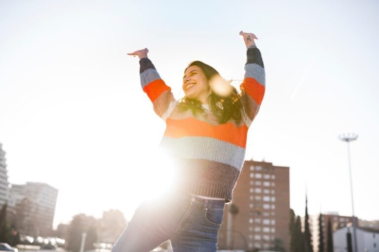 A young woman with long, dark hair, wearing a striped sweater and jeans, joyfully raises her arms in the air, surrounded by a sunny, urban scene.