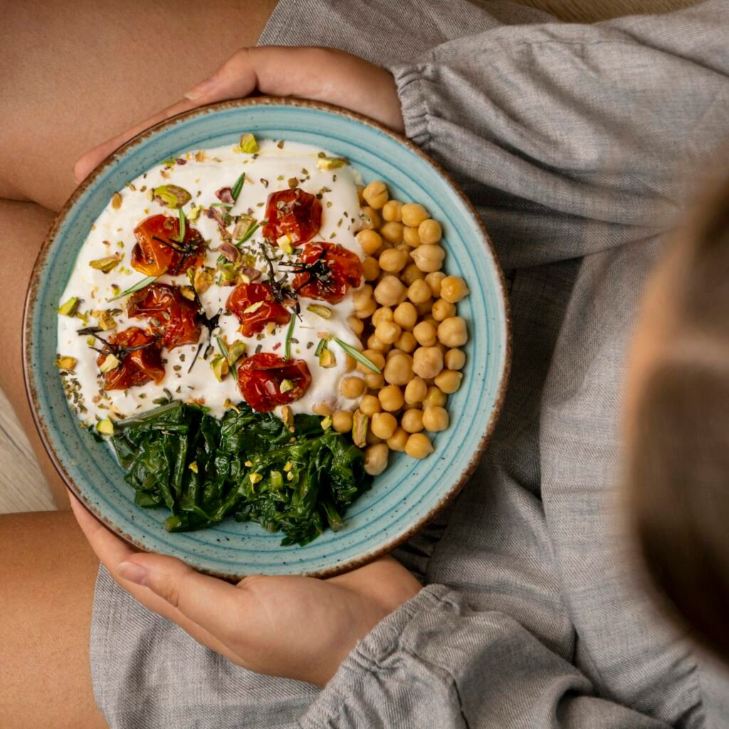 Overhead view of a girl's hands holding a bowl of healthy food: yogurt, chickpeas, spinach, and sun-dried tomatoes, staples of the Mediterranean diet. The girl wears a gray dress.