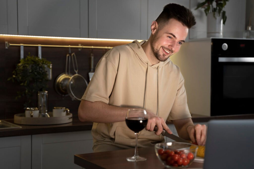 A man, smiling, prepares a meal in his kitchen, incorporating Temptation Bundling with the Mediterranean Diet by enjoying a glass of red wine while cooking.