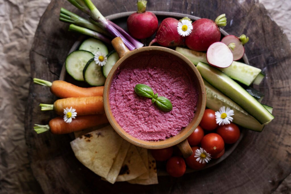 A vibrant overhead shot of a rustic wooden platter laden with colorful vegetables and a bowl of beet hummus, accompanied by pita bread.