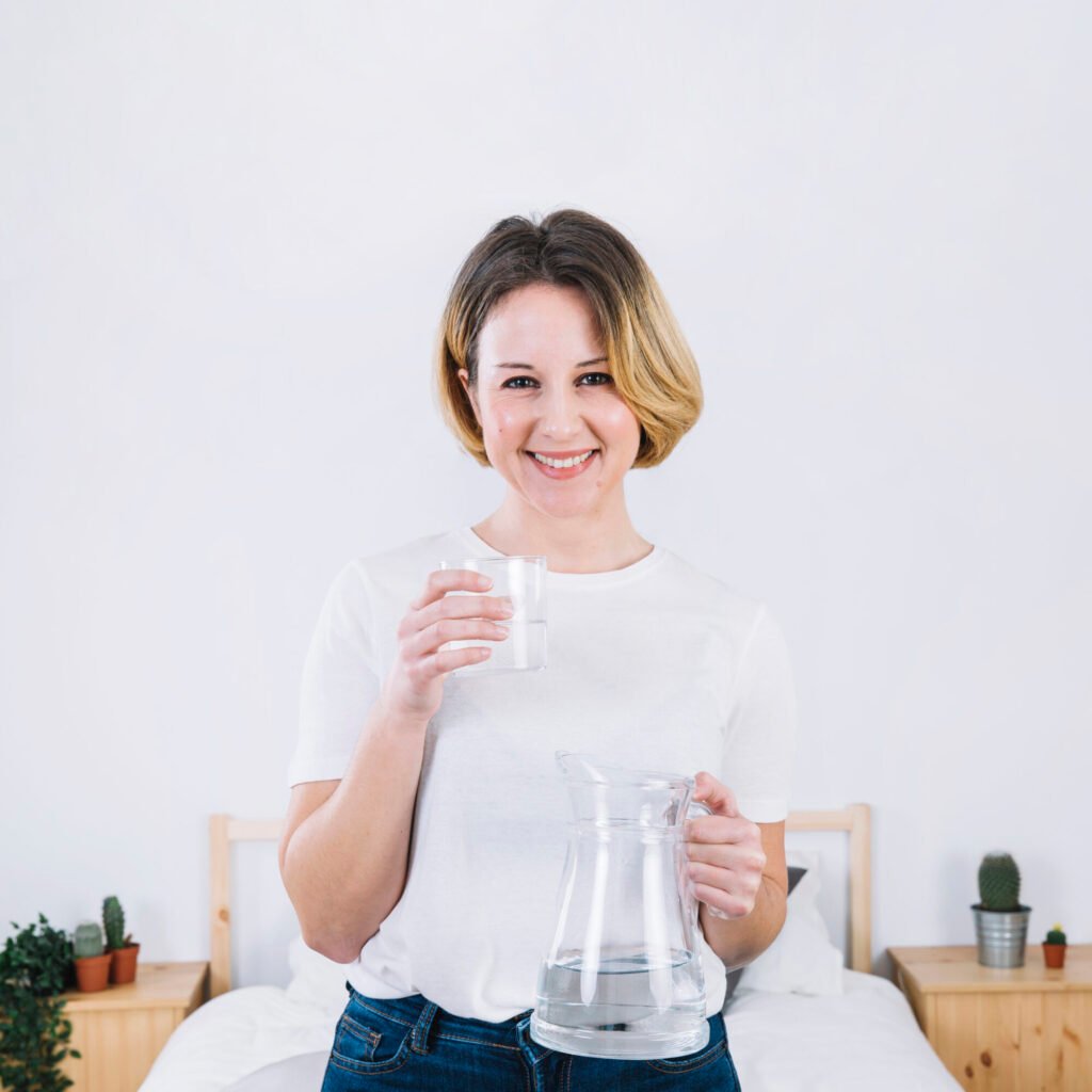 A smiling woman in a white shirt holds a glass of water and a water pitcher in a bedroom setting. She appears healthy and happy.

